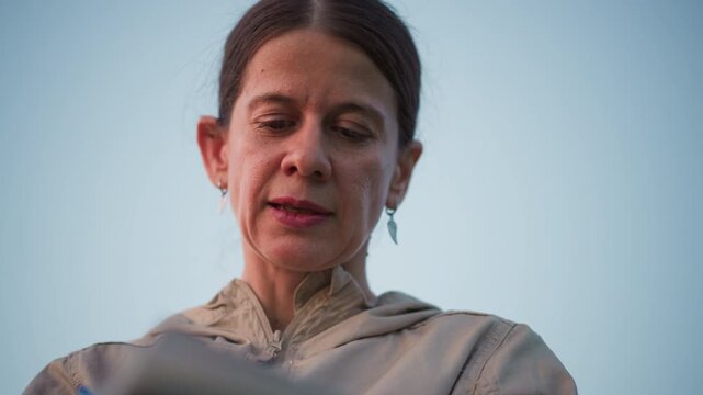 close up of middle age woman concentrating on writing in journal with calm focused expression outdoors against clear sky wearing beige jacket and leaf earrings
