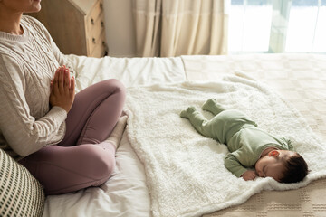 Mother is meditating beside sleeping infant on fluffy white blanket across quilt bed near window