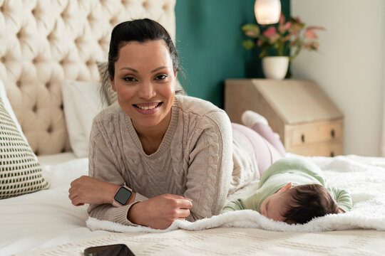 African American mother and baby reclining on bed under white blanket in bedroom beside smartphone
