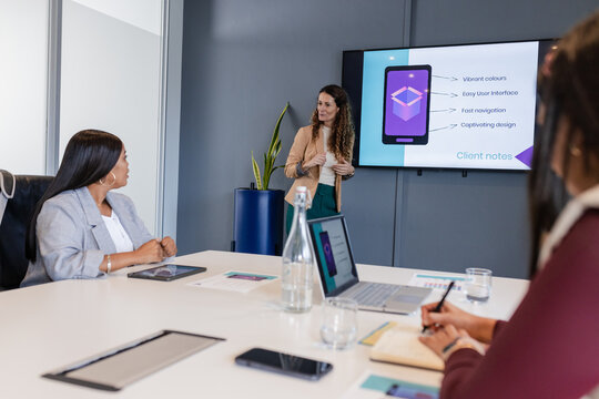 Diverse businesswomen presenting mobile interface slide at conference table with laptops tablets