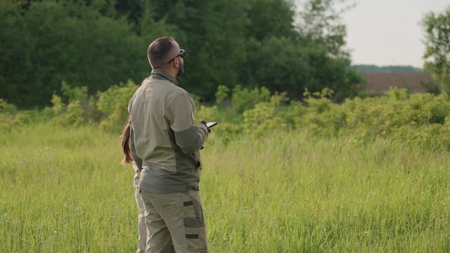 Researchers stand in tall grassy field holding device while scanning distant forest edge observing vegetation growth under bright summer daylight for scientific survey and data collection