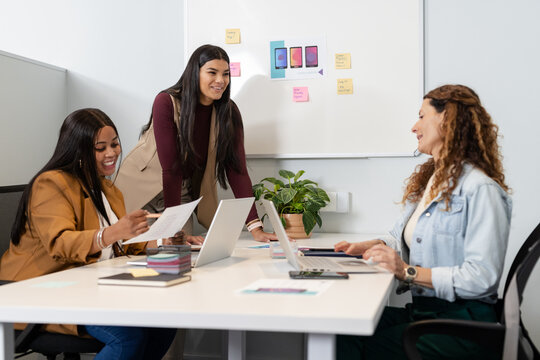 Diverse female colleagues collaborating at office table on app design with laptops and whiteboard