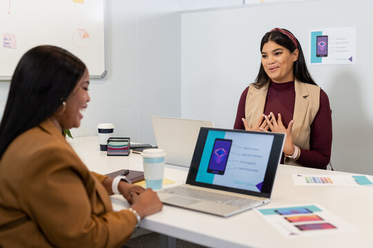 Diverse female coworkers discussing mobile app design at meeting room with laptops and mockups