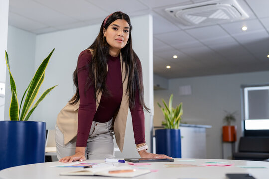 Woman in business attire leaning on white round table in office, sorting papers and sticky notes