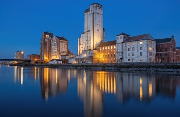 Obraz premium Serene cityscape river at dusk. Buildings reflect in calm water under blue sky. City lights glow warmly across urban landscape. River bank perspective offers panoramic view.