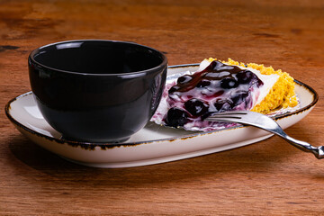 Side view of a cup of coffee in black ceramic cup with blueberry cheese pie on foil paper in white ceramic tray and metal fork on wooden table.