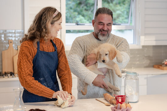 Couple kneading dough and holding golden retriever pup on marble countertop in kitchen - Powered by Adobe
