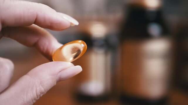 Close-up of fish oil capsule held by fingers. Soft light highlights golden hue. Ideal for health, wellness, and supplement-related content.