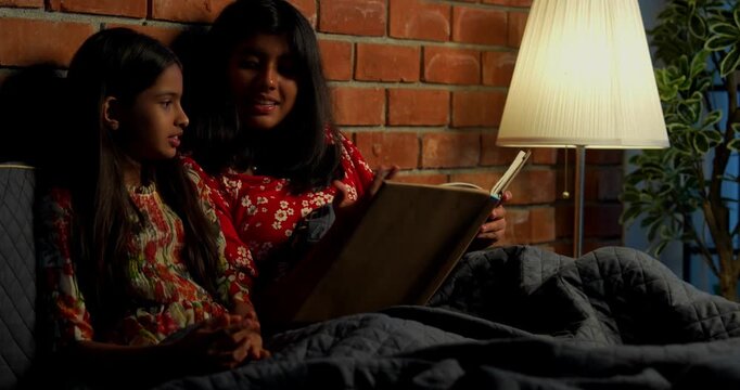 Indian girls reading bed time stories before falling asleep under cozy quilt at night. Asian little sibling sisters lying together in bed, enjoying nighttime routine in warm indoor bedroom setting