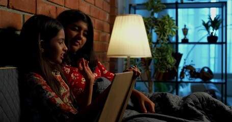 Indian girls reading bed time stories before falling asleep under cozy quilt at night. Asian little sibling sisters lying together in bed, enjoying nighttime routine in warm indoor bedroom setting - Powered by Adobe
