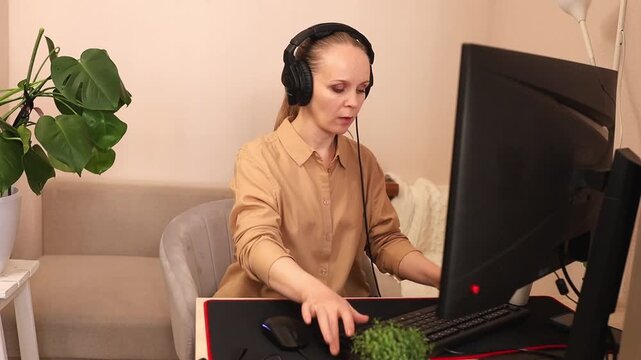 A focused young white woman is working on a computer at her desk. She's wearing headphones. Listening to music. Watching an online lesson.