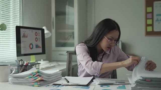 Stressed young businesswoman struggling with overwhelming paperwork, experiencing workplace tension while analyzing documents at office desk