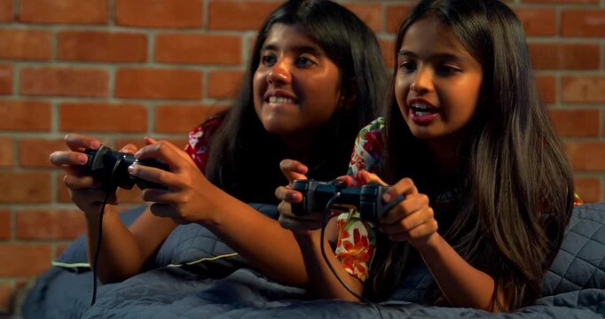 Indian sibling sisters playing video game lying on bed and enjoying fun time together. Asian little girls holding gaming controller while relaxing indoors in cozy bedroom environment with happy faces