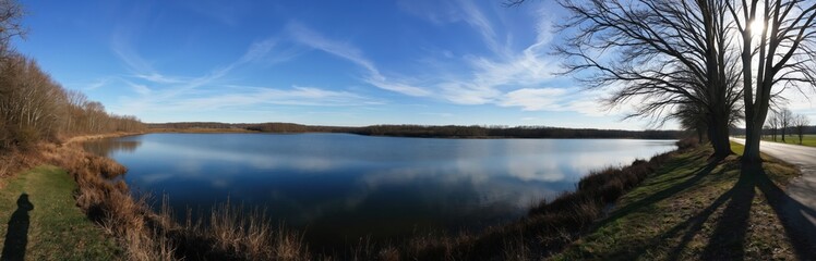 Fototapeta premium Panoramic view of tranquil lake reflecting clear blue sky with wispy clouds. Grassy area and trees line water edge. Road runs parallel to lake, bordered by tall trees casting long shadows.