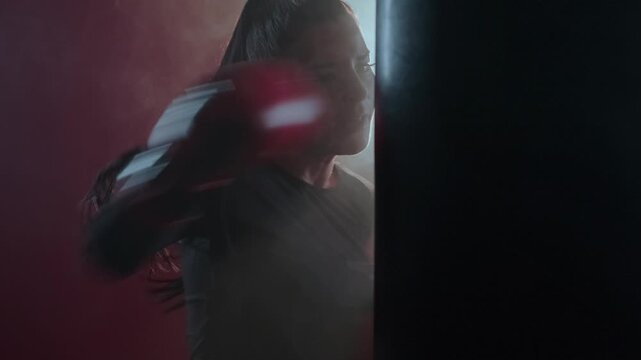 Slow motion shot of a female fighter punching a heavy bag in the gym while wearing boxing gloves. Intense focus and strength captured in dramatic lighting, showcasing power and precision. 4k footage.
