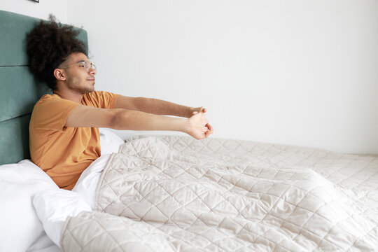 Side view indoor image of african american young male tourist waking up in his hotel room bed, stretching his hands sitting in front of window, planning to enjoy strolling in new place