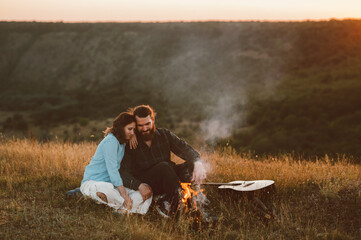 A cozy couple is enjoying a warm campfire together during a serene sunset while surrounded by...
