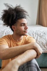 Young sad pensive black man being depressed having troubles in his relationships, sitting alone on floor leaning his back against bed and looking aside through window, ready to burst in tears