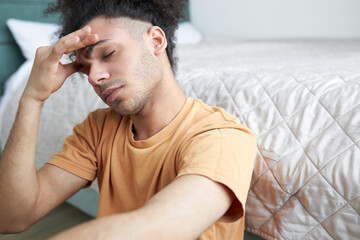 Young man in orange shirt holding his forehead with closed eyes, suffering migraine or headache, sitting on floor in his bedroom with closed eyes, feeling unwell after waking and getting up