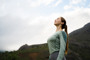 Young woman in sportswear breathing in fresh air outdoors, girl with closed eyes enjoying nature while meditating, standing in mountains on cloudy day, mindfulness lifestyle, low-angle view