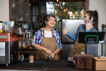 Older and younger woman baristas working together behind the counter, smiling and talking, showing happiness, teamwork, and connection in a small coffee shop business.
