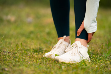 Close-up of woman in sportswear holding ankle in pain after outdoor workout, injury or muscle strain during fitness activity in nature, consequences of overtraining concept