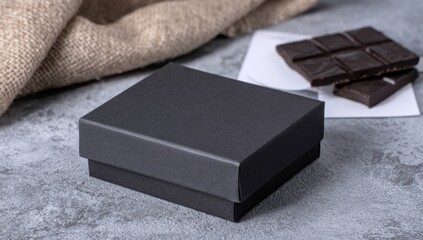 Dark chocolate pieces rest by a small, plain black box, a burlap sack in the background, all on a textured gray surface