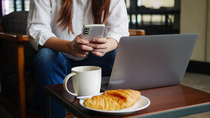 front view of woman using phone and laptop computer at home or coffee shop for work in the morning with hot coffee, anonymous face. working from home and remote work concept