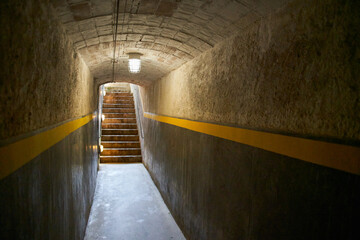 Interior of a narrow anti-aircraft tunnel with a light bulb on the ceiling and at the end some illuminated stairs leading up with a yellow line painted on the walls