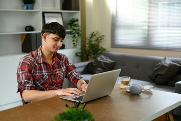 Young man in a casual red plaid shirt working on a laptop at home with coffee on the table