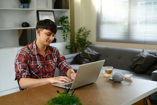 Young man in a casual red plaid shirt working on a laptop at home with coffee on the table