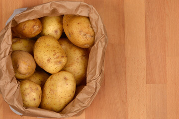 A brown paper bag filled with fresh, unwashed yellow potatoes sits on a wooden surface from a top-down perspective with ample copy space on the right