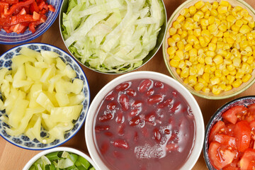 An overhead flat lay captures a vibrant array of fresh, colorful chopped salad ingredients, including red and yellow bell peppers, lettuce, corn, kidney beans, and tomatoes, arranged in various bowls 