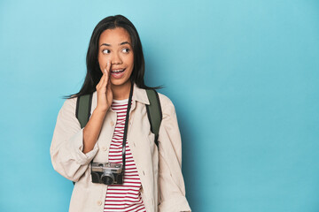 Filipina woman with camera and backpack on blue is saying a secret hot braking news and looking...
