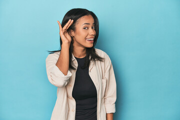Filipina young woman on blue studio trying to listening a gossip.