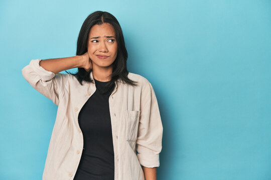 Filipina young woman on blue studio touching back of head, thinking and making a choice.