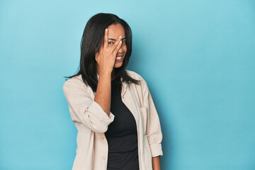 Filipina young woman on blue studio blink at the camera through fingers, embarrassed covering face.