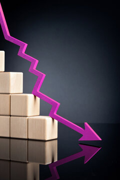 A purple downward zigzag arrow next to with stacked wooden blocks, symbolizing a declining trend on a reflective dark surface. High quality photo.
