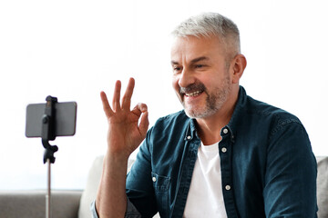 Smiling handsome senior man vlogger broadcasting from home, sitting on sofa and showing okay gesture at mobile phone camera. Male elderly blogger having stream, chatting with followers, copy space