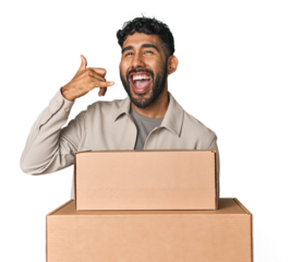 Hispanic male holding cardboard boxes in studio showing a mobile phone call gesture with fingers.