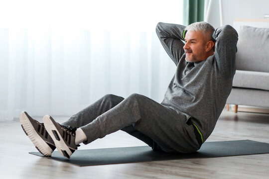 Matured man in grey sportswear training at home, laying on fitness mat and doing crunches, working on his abs musculs, using modern smartphone on tripod, watching online fitness lessons, free space