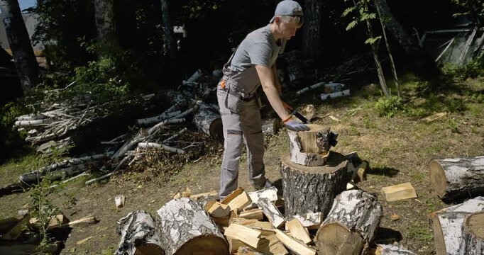 Man Splitting Large Birch Logs with Axe Outdoors in Sunny Forest Area, Chopping Firewood for Winter, Dynamic Action Scene with Wood Chips Flying, Stack of Birch Firewood and Tree Trunks in Background