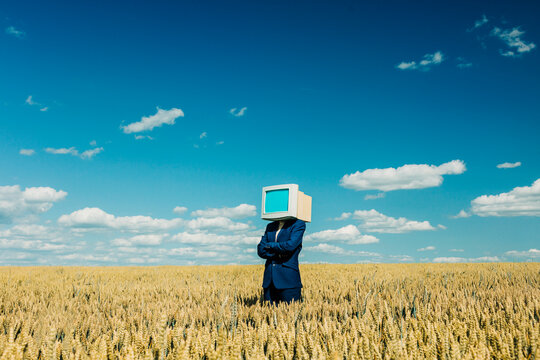 Businesswoman with retro monitor head stands in wheat field under blue sky