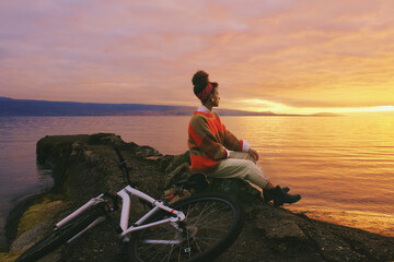 Outdoor portrait of happy african woman admiring sunset over the lake during bike ride