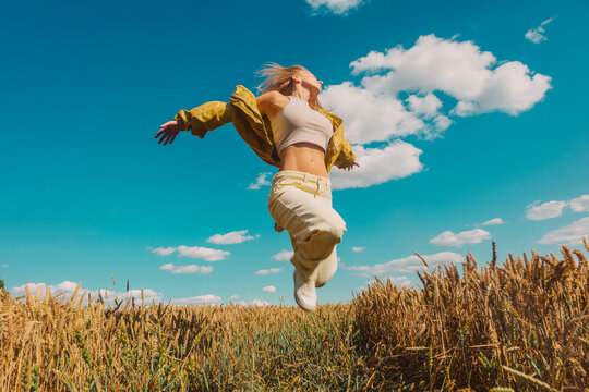 Stylish woman with blonde hair jumping in wheat field under blue sky