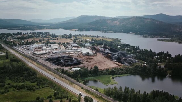 Aerial timelapse video of Priest River, Idaho, shows an industrial lumber yard with stacked timber piles beside the river, mountains in the background, and a highway cutting through the green valley.