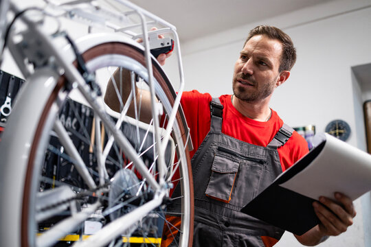 Experienced serviceman holding checklist and inspecting bicycle inside workshop.