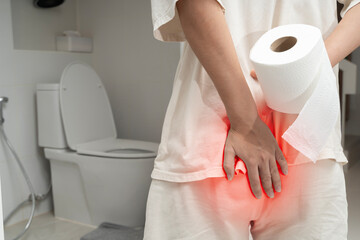 Person holding toilet paper and touching painful buttocks area with red highlight in front of toilet seat