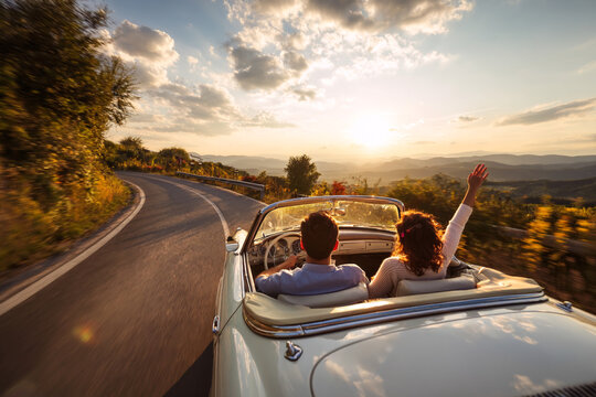 Couple driving a vintage convertible car on a winding road at sunset with one person raising their hand joyfully.