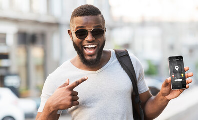 Excited black guy pointing at mobile taxi application on his phone while waiting for cab on city street, panorama. African American male passenger promoting online car booking service